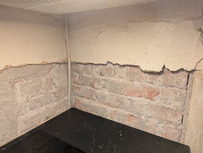 Renoveringshörn i kök med blottade tegelstenar, avskalad puts, svart bänkskiva och en synlig kabel.