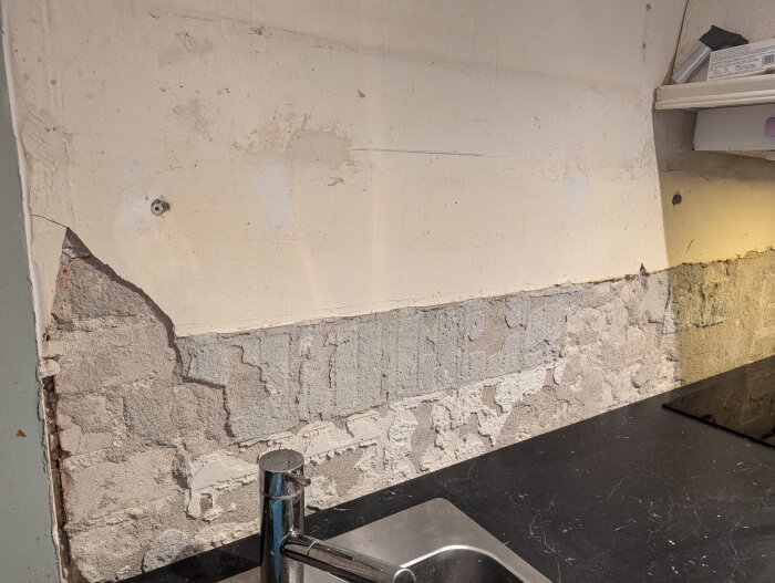 Sliten puts på en köksvägg ovanför en svart bänkskiva, med synlig sprucken yta och en kromfärgad kran, under renovering.
