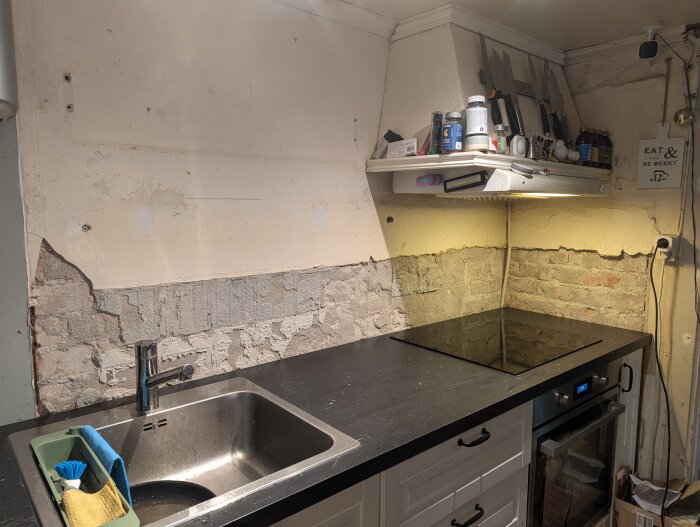 Kök med svart bänkskiva, diskho, och spishäll. Vägg med borttagen kakel och synlig puts ovanför bänkskivan. Hylla över spisen med köksredskap.