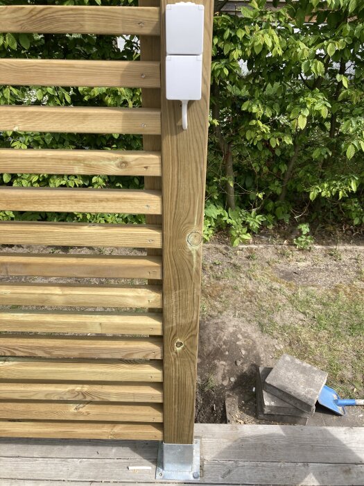 Trästaket med horisontella ribbor och en väggmonterad eluttag, omgiven av gröna buskar och stenplattor på marken.