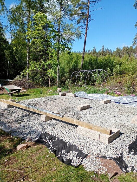 Grund till friggebod med markduk, sättsand, makadam och lecablock i trädgårdsmiljö. Träd och växtlighet i bakgrunden.