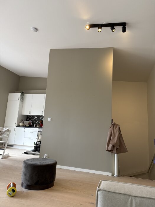 Inomhusvägg i ett modernt vardagsrum, beige vägg med plankgolv, spotlight i taket, puff och leksak på golvet. Köksdel med vita skåp syns i bakgrunden.