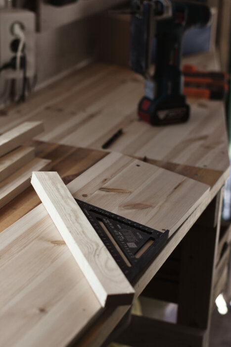 Arbetsbänk med trälister, vinkelhake och skruvdragare i bakgrunden, del av hemmagjorda sågskenor.