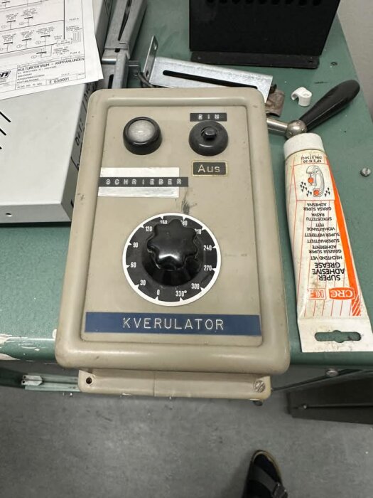 En "Kverulator" med vridreglage och knappar, placerad bredvid super adhesive grease i en verkstadsmiljö.