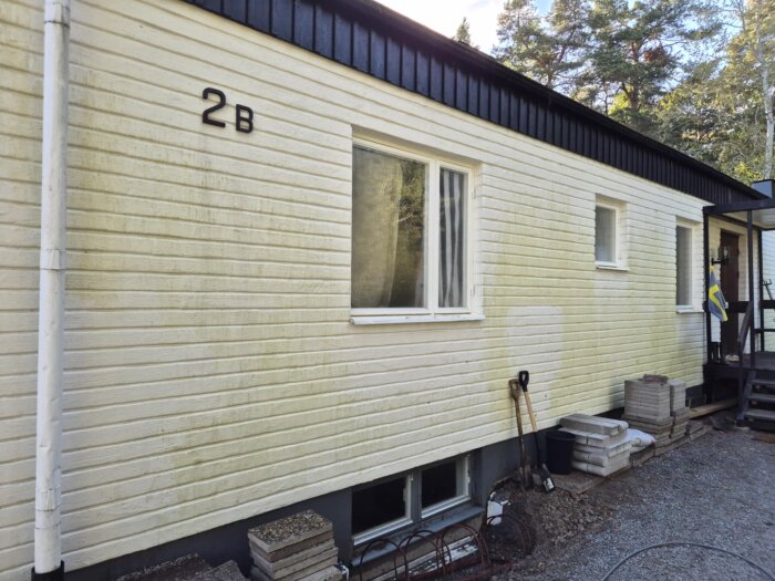 Gulnad träfasad på ett hus med viss algpåväxt, sedd före fasadtvätt. Grusmark med staplade plattor och verktyg lutade mot väggen.