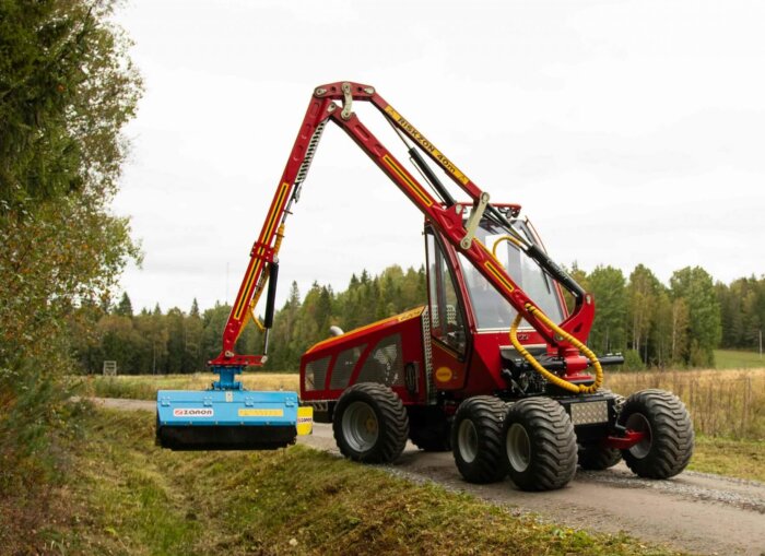 Röd skogsmaskin med kran och blå klippaggregat, används för landsskapsvård. Omgiven av skogslandskap.