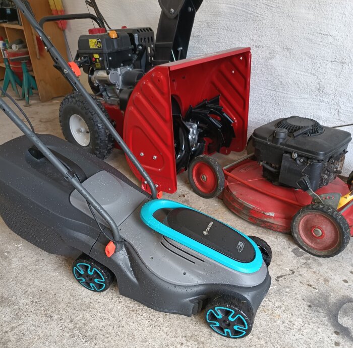 Gardena 18v gräsklippare med blå detaljer i ett garage bredvid en röd snöslunga och en röd äldre gräsklippare.