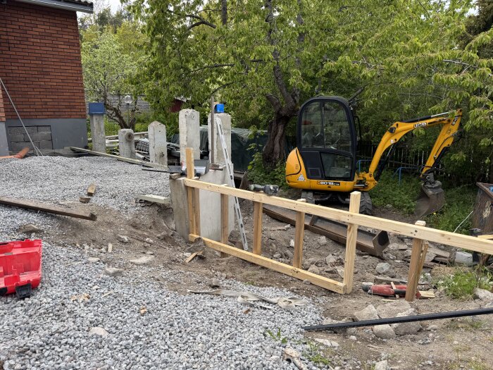 En byggarbetsplats med stödmurar under konstruktion, en grävskopa och en byggnad i bakgrunden, omgiven av grönska.