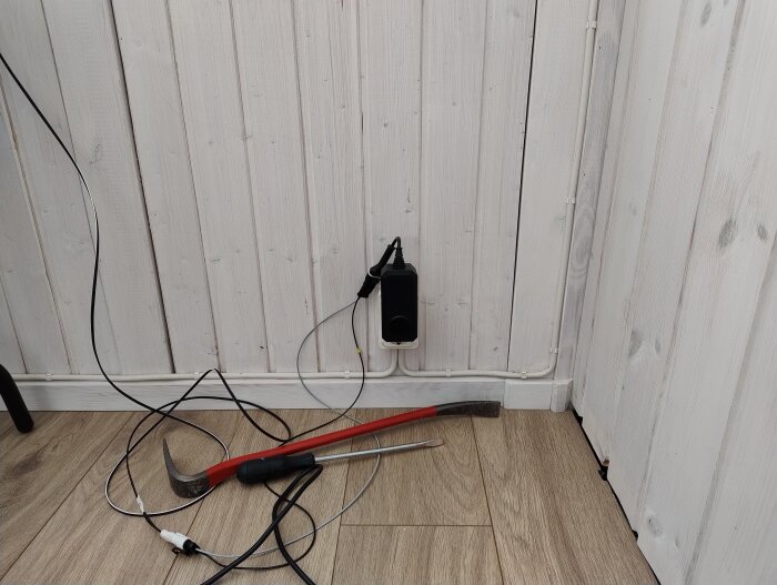 Vägguttag med sladdar och verktyg på ett trägolv; kablar löper längs väggen i anslutning till altan.