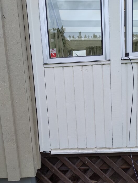 Utsida av ett hus med träpanel, ett fönster och en svart kabel som går ut genom fönstret.