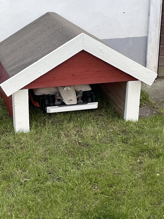 Rött litet träskjul byggt av spillvirke, huset är målat med vit och röd färg, en robotgräsklippare står under taket på en gräsmatta.