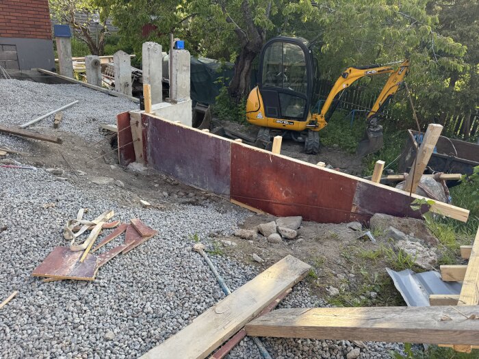 Byggarbetsplats med grävskopa och pågående gjutning av armerad stödmur av träform vid kant av stenläggning i en trädgård.