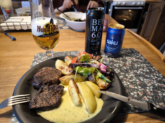 Grillad filé med klyftpotatis, sallad och sås på svart tallrik, öl i Hard Rock-glas och två ölburkar på bord med blommig underlägg.