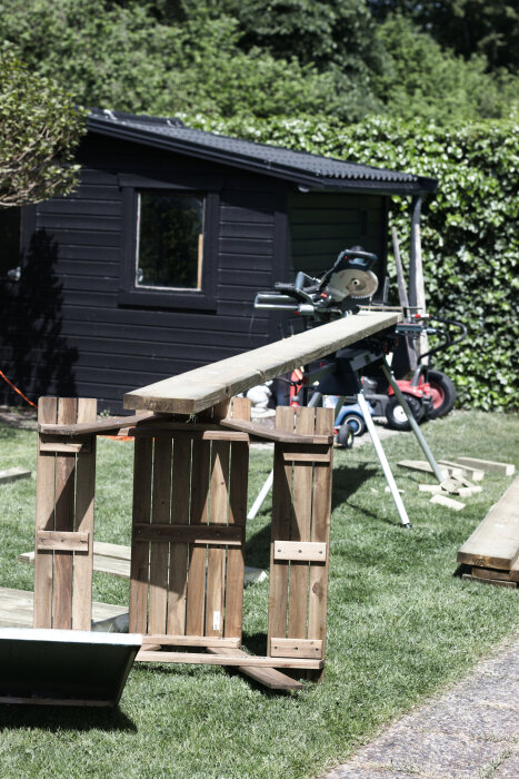 En arbetsplats utomhus med kap- och gersåg på ställning. En träregel vilar på två upprättstående lastpallar framför ett svart skjul.