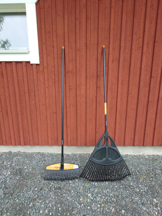 Två lövkrattor, en med platt huvud och en solfjäderformad, står lutade mot en röd trävägg.