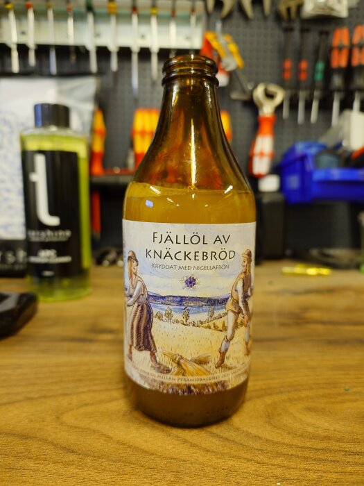 Flaska med etiketten "Fjällöl av Knäckebröd", kryddat med nigellafrön, står på en arbetsbänk i en verkstad.