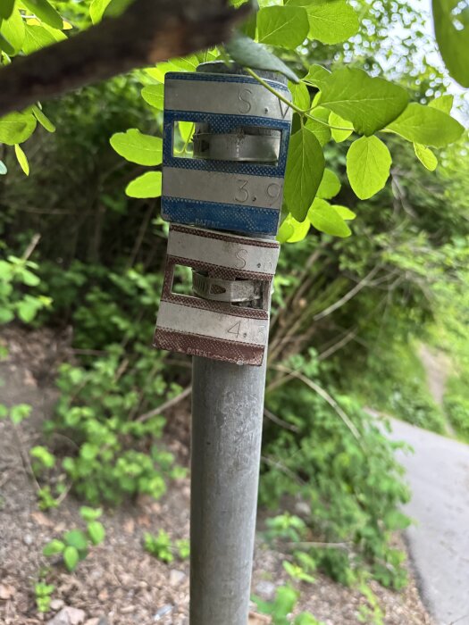 Metallstång med två färgade skyltar i skogsområde, med numrering och bokstäver. Möjligen markering av en tomtgräns.