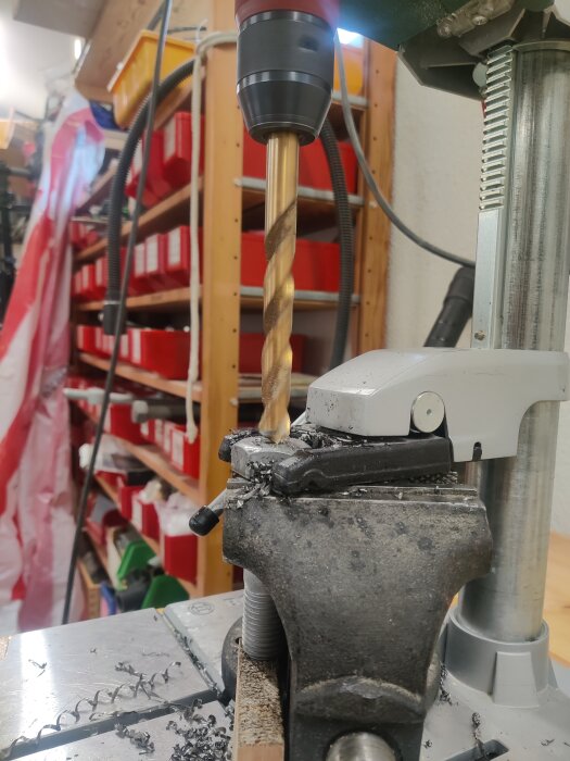 Borrmaskin borrar hål i metallbult fastspänd i skruvstäd på arbetsbänk, hyllor i bakgrunden.