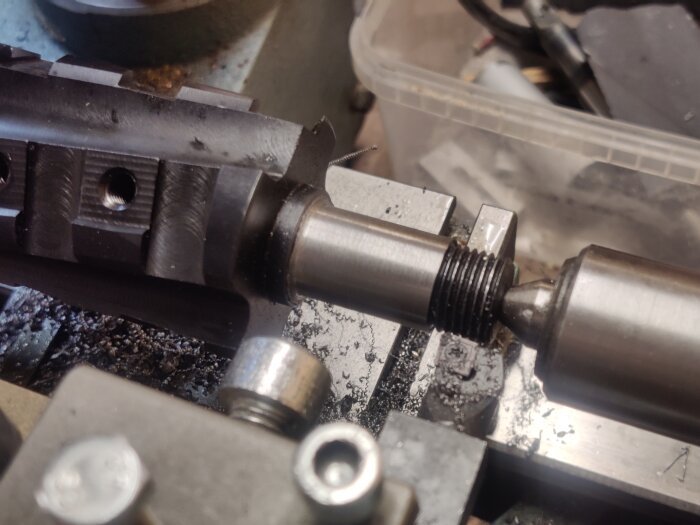 Närbild på en svarvmaskin som bearbetar en metallstång med gängor, omgiven av metallspån och verktygsdelar.
