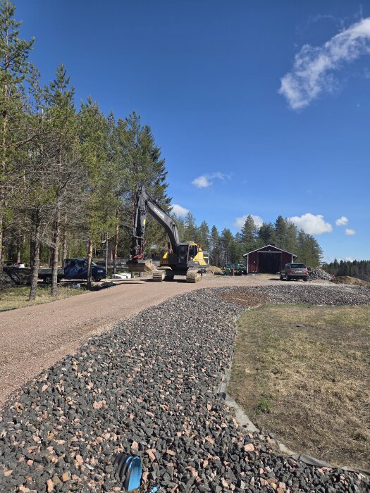 Grävmaskin på grusväg intill skog, upplagd marksten i förgrunden, blå himmel i bakgrunden.