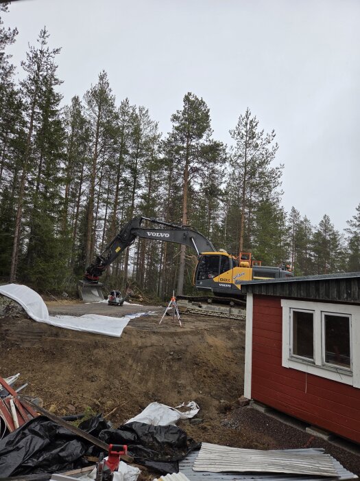 Grävmaskin på kulle vid skogsbrynet, omgiven av träd och en röd byggnad i förgrunden.
