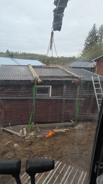 Lyft av bastutak med grävskopa och kedjor, placeras på ny plats på kulle. I förgrunden syns en liten eld och byggmaterial.