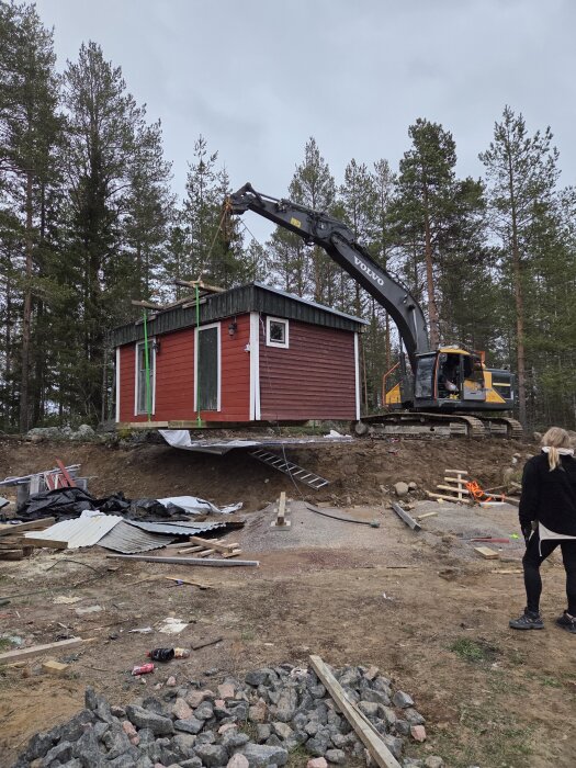 Grävmaskin lyfter röd bastustuga till en ny plats på en kulle i en skogsmiljö. Byggmaterial och verktyg syns på marken.