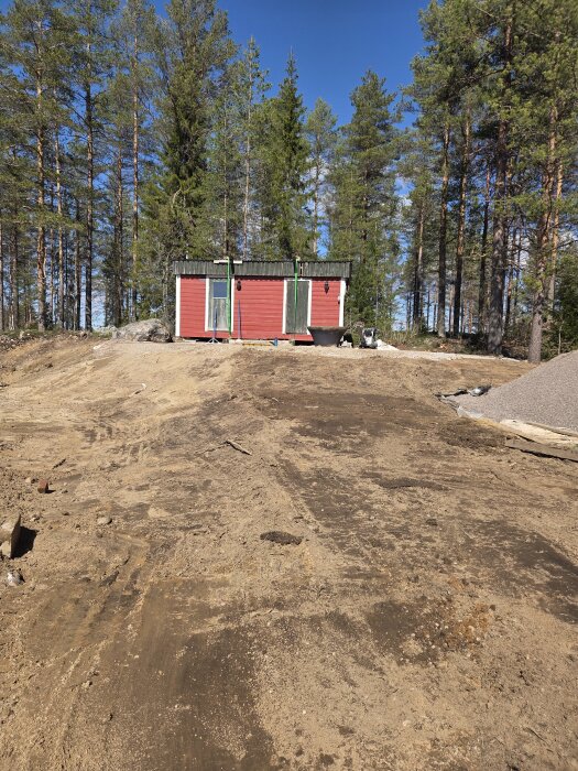 Röd bastubyggnad placerad på en ny kulle, omgiven av skog. Marken runtomkring visar tecken på schaktning och förberedelse för vidare byggen.