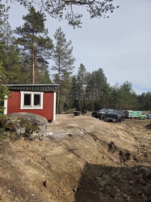 Röd bastubyggnad på en kulle, omgiven av grävd mark och en parkerad SUV, med skog i bakgrunden.