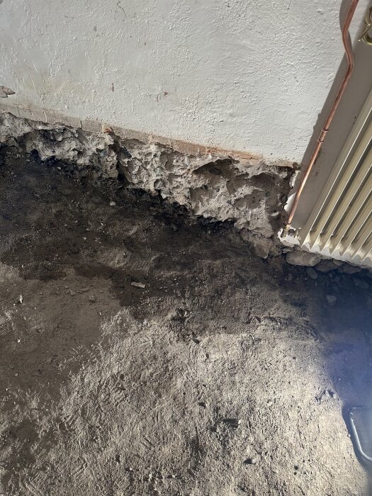 Nyligen bilat källargolv visar fukt mot en ojämn vägg med synlig spricka och radiatorrör i ett hus byggt 1911.