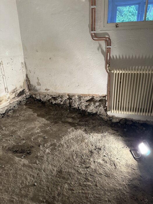 Källarrum med bilat betonggolv, exponerade rör på väggen, radiator och ett fönster. Golvet visar tecken på fukt och sprickor nära väggen.