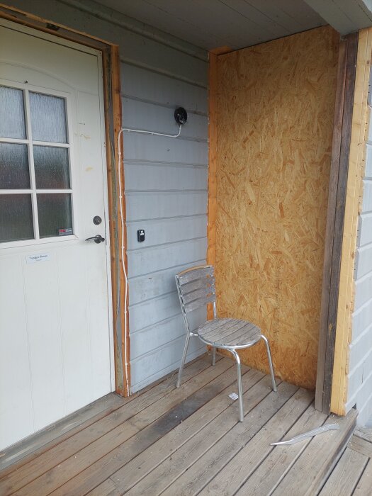 En altan med en vit ytterdörr, träpanelvägg, plywoodskiva, och en metallstol står på trägolv; del av en renovering.