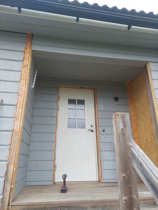 Entré till hus under renovering med sliten träpanel och vit dörr, elverktyg på verandagolv.