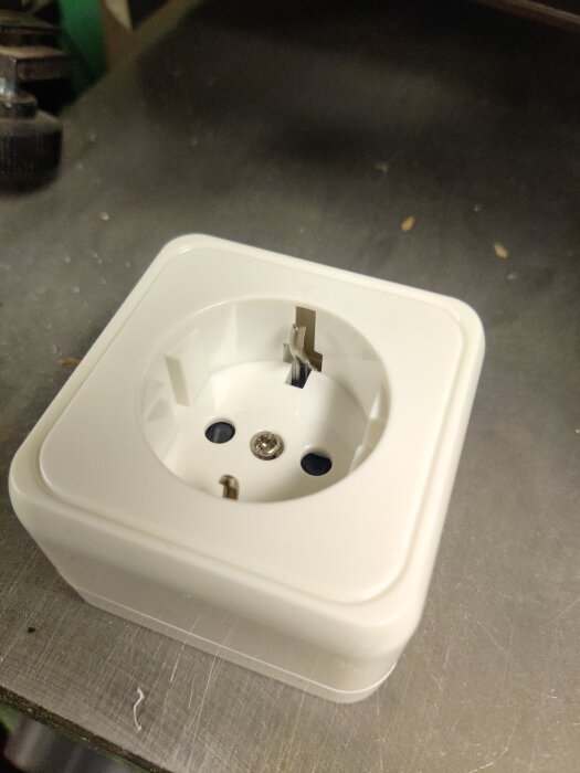 Vitt eluttag med skruvanslutning och metallbleck, kritiserat för dålig design och kvalitet enligt inlägg. Placeras på en metallisk yta.