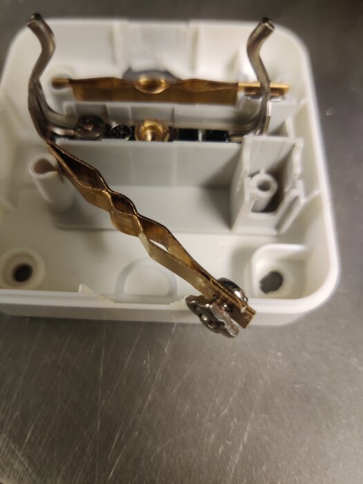 Trasigt eluttag med rackliga skruvanslutningar och metallbleck instuckna mellan plastflärpar, delvis demonterat, liggande på ett bord.