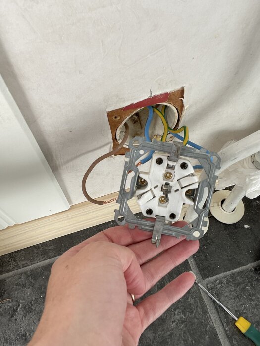 Eluttag med lösa kablar, brun och blå borttagna, hand håller uttag med vit plastdel och verktyg på golvet.