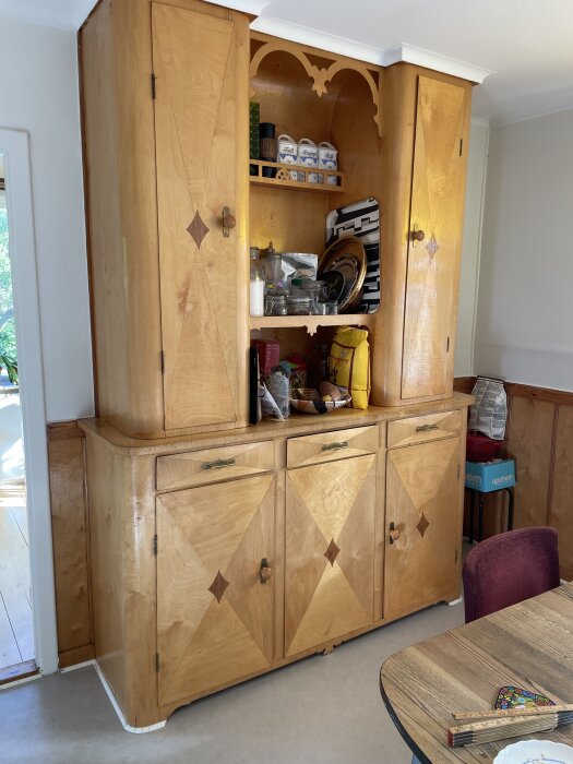 Köksskåp i trä från 1939 med dekorativa sniderier, tvådelad design med hyllor fyllda med köksartiklar och dörrar med triangulära mönster.