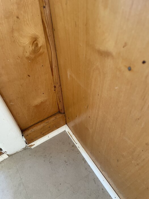 Hörn av ett träköksskåp från 1939 fast i en vägg, med sockel och golv av linoleum. Skador syns på sockeln i skarven.