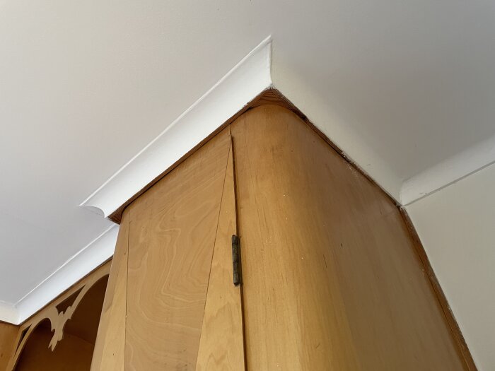 Köksskåp av trä, fäst mot en vit vägg och tak, med synliga gångjärn och dekorativa detaljer.