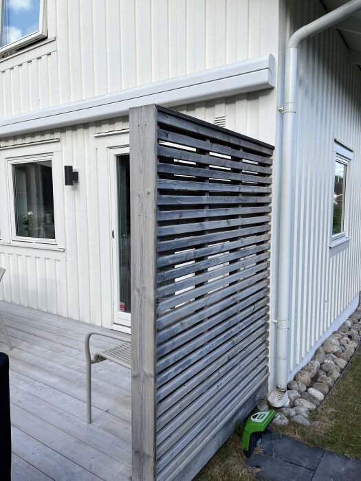 Husvägg med vit träpanel, trätrall och en spaljé; stuprör och stenar längs grunden. En lampa och markis ovanför fönstren.