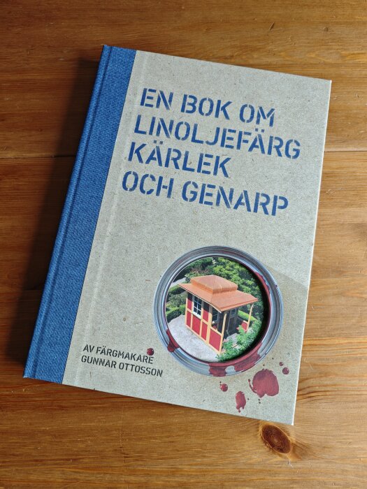 Bok med blå rygg på träbord. Titeln "En bok om linoljefärg, kärlek och Genarp" och bild på litet hus i färg. Av färgmakare Gunnar Ottosson.