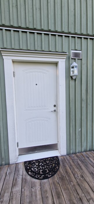 Vit dörr på grön fasad med frånluftsvärmepumpens utblås ovanför dörren bredvid en utomhuslampa och en svart dörrmatta på altangolvet.