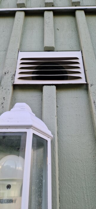 Utblås från frånluftsvärmepump på husfasad med vita lameller och en vit utomhuslampa nedanför.