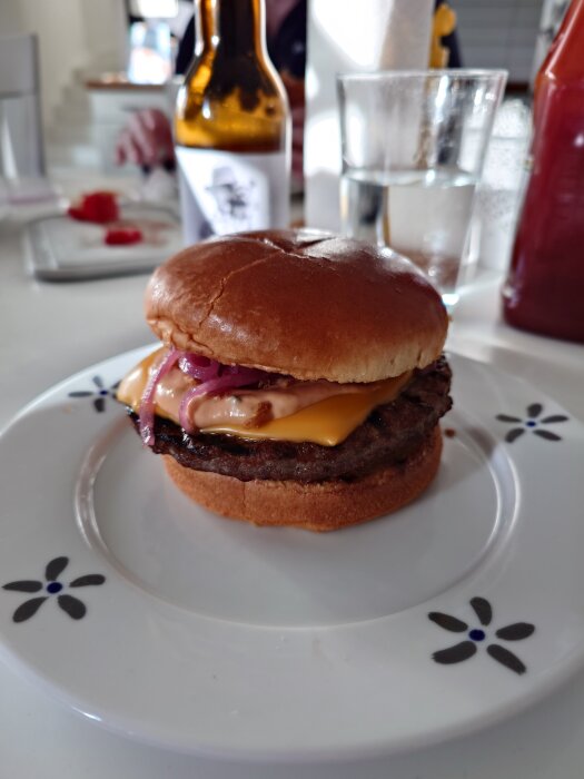 Hamburgare på tallrik med ost, lök, burgardressing, senap och ketchup. I bakgrunden syns en glasflaska och vattenglas.