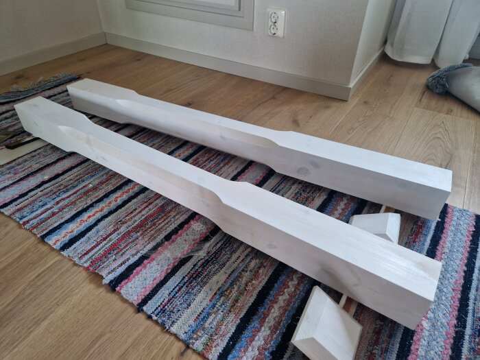 Två vita trästolpar ligger på en randig matta på ett trägolv, redo att monteras för en trappa.