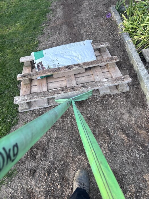 Person drar en lastpall med säckar över jord på en grusgång, grönt band används, trädgårdsarbete pågår nära gräsmatta och blomsterbänk.