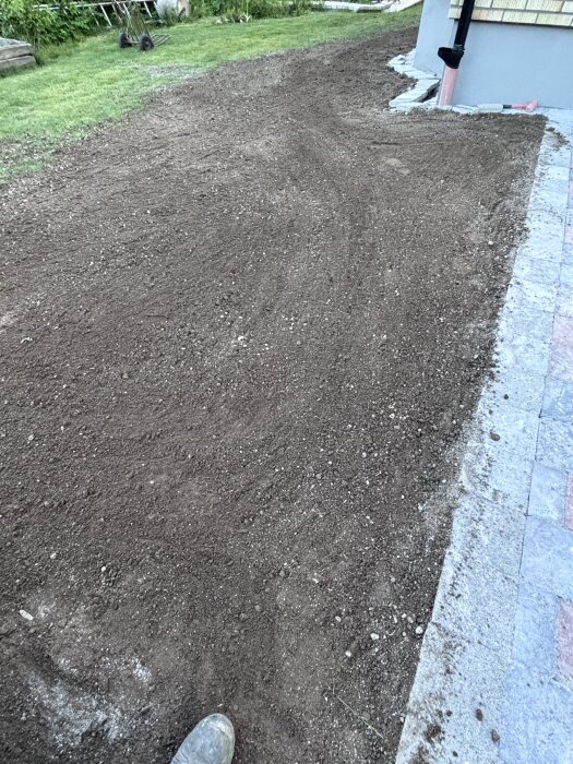 Nyjordad mark bredvid stenhällor, redo för gräsplantering. Margaret är lätt synlig, antyder arbete med att packa jorden.