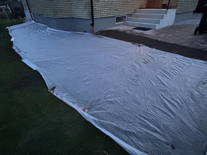 Gräsmatta täckt med presenning nära husvägg, del av renoveringsprojekt för att fylla ut ytan med ny jord och så gräs.