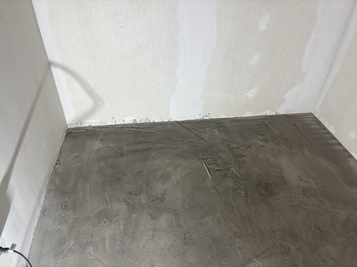 Ett nyflytspacklat golv i ett rum där väggen visar tecken på pågående renovering. Golvets yta är ojämn och skiftande i struktur.