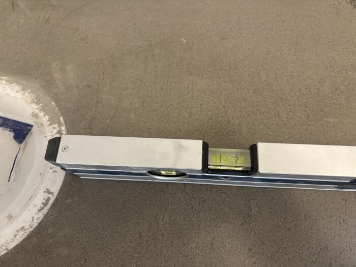 Vattenpass placerat på golv med flytspackel intill en avloppsbrunn för att kontrollera lutningen.